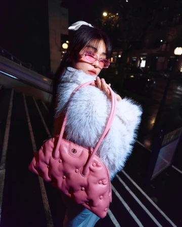 A woman in a fluffy white jacket and pink tinted glasses holding a pink leather handbag on outdoor stairs at night.